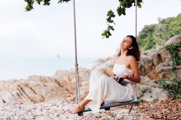 Romantic young happy calm caucasian tattooed lonely woman with ukulele on swing tropical rocky beach at sunset   - Фото, изображение