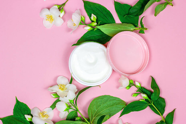 Top view of white plastic cosmetic container for moisturizing cream as a mock up with bright fresh jasmine flowers on pink background with copy space. Body and skin care and beauty concept. - Photo, Image