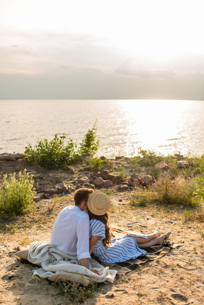 back view of young woman in straw hat and man sitting on blanket near river  - Foto, Imagem