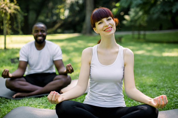 Front view of multiethnical couple practicing yoga in the park. Smiling pretty Caucasian woman sitting in lotus pose on the mat. Cheerful African man is happy sitting behind woman - Fotoğraf, Görsel