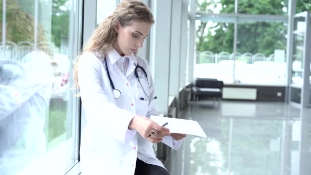 Smiling female doctor with a folder in uniform sitting. Attractive doctor in white coat and stethoscope smiling at camera in clinic - Imágenes, Vídeo