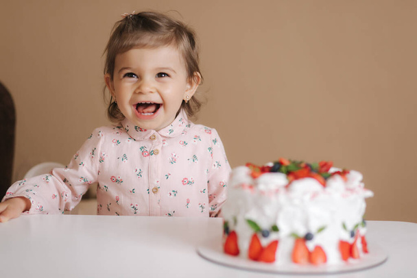 Χαριτωμένο κοριτσάκι ένα και ένα Χαλφ-year old σταθεί από νόστιμα τούρτα γενεθλίων. Δεκαοχτώ μηνών κορίτσι πολύ χαρούμενο και γελά - Φωτογραφία, εικόνα