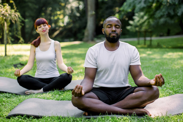 Close-up of multiracial couple taking a rest in the park, practising yoga, sitting in lotus position on yoga mats with hands in Zen, deep breathing, eyes closed. Focus on African man - Photo, image