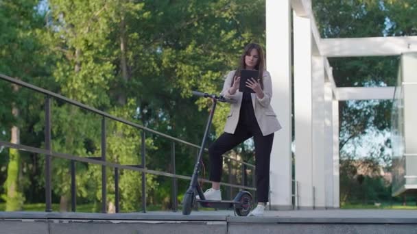 Una giovane bella donna è in piedi accanto al suo scooter elettrico, con un piede su una piattaforma in piedi. Sorride, è preparata per gli affari, con un'architettura moderna sullo sfondo. - Filmati, video