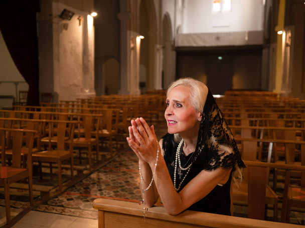 Olgun bir kadının yatay portresi. Kilisede oturmuş tespihle dua ediyor. - Fotoğraf, Görsel