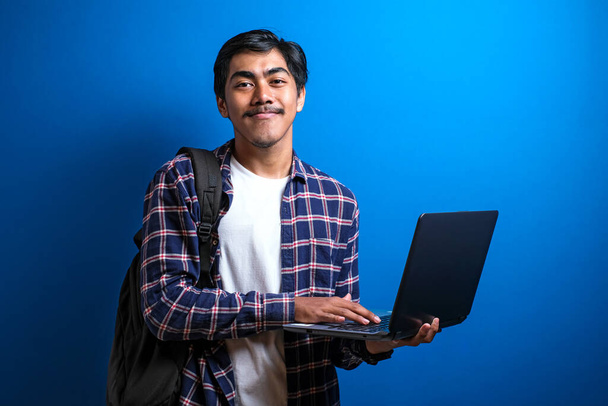 Sonriente joven asiática estudiante universitario usar camisa celebrar moderno portátil contra bluebackground con espacio de copia
. - Foto, Imagen