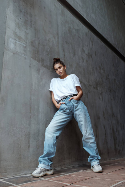 A beautiful young woman in full growth stands against the wall. A stylish girl in blue jeans and a white T-shirt stands against the background of a gray concrete wall. 80's 90's style - Valokuva, kuva