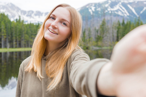 Η τουρίστρια που βγάζει σέλφι στη λίμνη του βουνού. Κοιτάζοντας την κάμερα και χαμογελώντας. Ταξίδι και ενεργή ζωή έννοια. Εξωτερικές πόρτες - Φωτογραφία, εικόνα