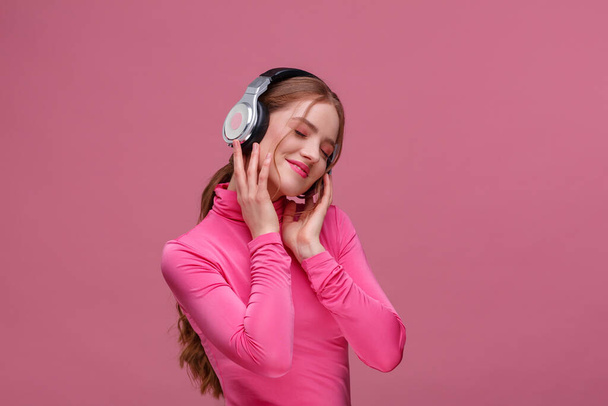 音楽を聴いてお楽しみください。ヘッドフォンの音楽を聴いて美しい若い赤毛の女性。イヤホンの面白い笑顔の女の子とピンクの背景にダンスピンクのブラウス。リラクゼーションとストレス管理 - 写真・画像