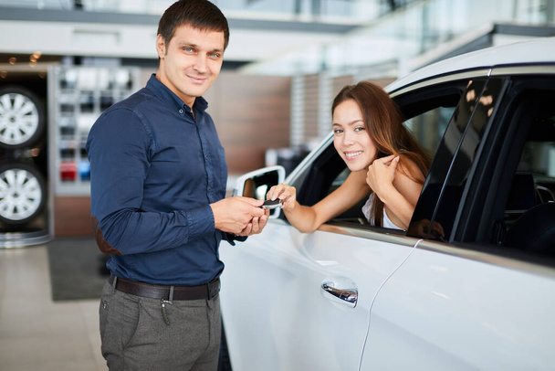 ar salesman giving car keys to young woman sitting in car in auto dealership - Photo, image
