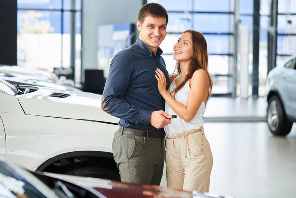 Happy loving couple embracing near their new luxury car at dealership. - Photo, image