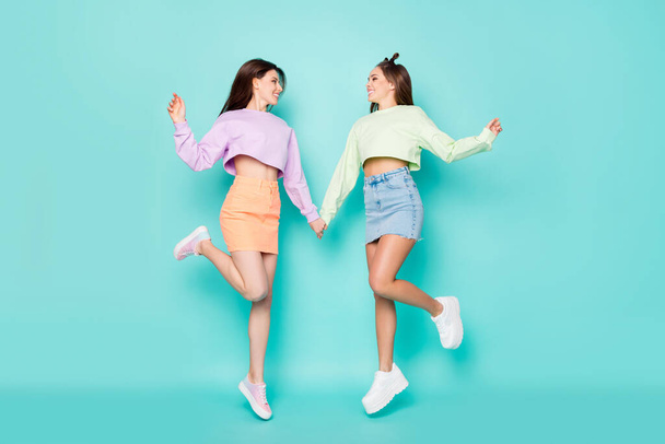 Full length photo of two pretty crazy lady sisters jump high cool youth clothes hold arms look eyes wear cropped sweaters short skirts shoes isolated pastel teal color background - Foto, afbeelding