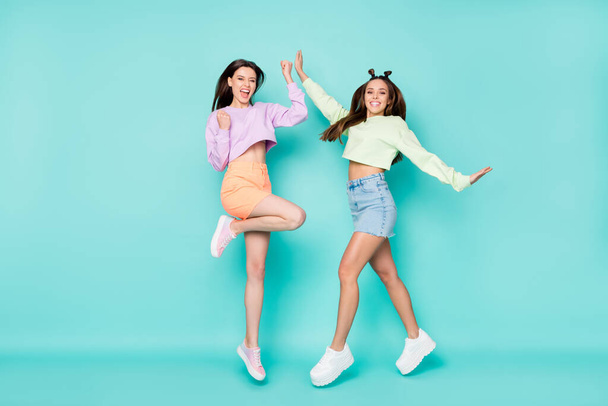Full size photo of two pretty crazy lady sisters jump high up youth clothes rejoicing students party wear cropped sweaters short skirts shoes isolated pastel teal color background - 写真・画像