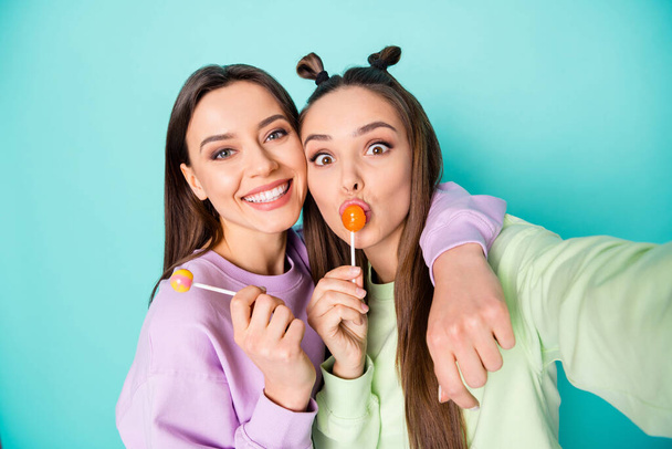 Photo of two funny ladies hold lollipop chupa chups hands childish tasty sweets making selfies wear green violet pullovers isolated pastel teal color background - Φωτογραφία, εικόνα