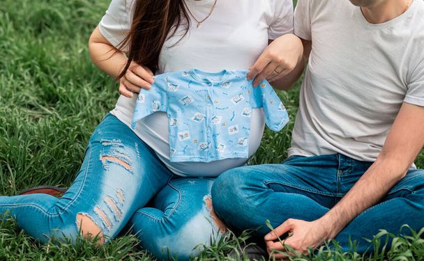 Imagem cortada da mulher grávida segurando roupas de bebê com o marido no campo verde. Os momentos ternos sinceros. - Foto, Imagem