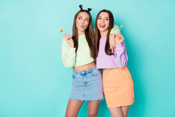 Photo of two funny lady hold lollipop chupa chups sweets addicted send kiss lick lips wear cropped pullovers naked belly mini skirts isolated pastel teal color background - Фото, изображение