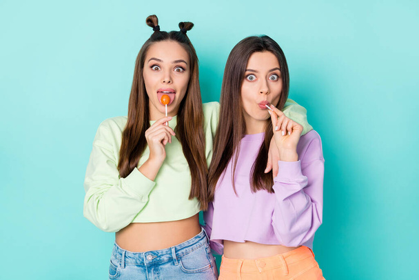 Photo of two crazy funny cheerful ladies hold lollipop chupa chups hands childish mood tasty sweets wear cropped pullovers naked belly skirts isolated pastel teal color background - Фото, изображение