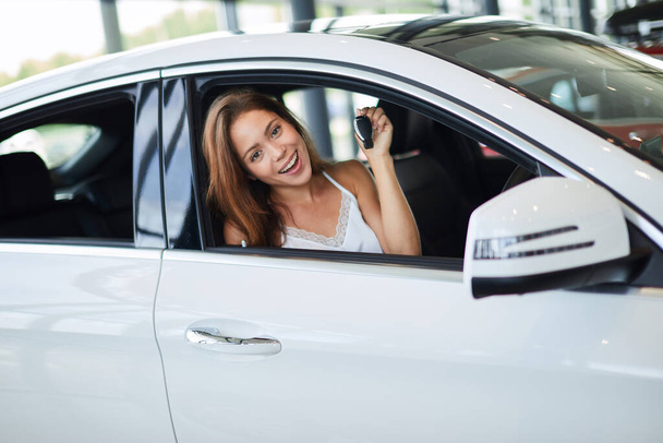 Happy woman driver in white car holds car keys in her new car at car show room - 写真・画像