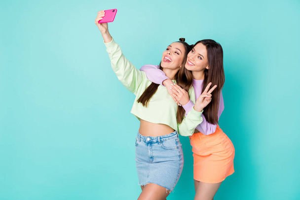 Photo of two pretty cheerful ladies best friends hold telephone take selfies showing v-sign symbol wear cropped sweaters naked belly short skirts isolated pastel teal color background - 写真・画像