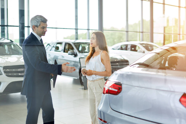 Young female customer asking mature sales manager to help in car dealership. - Foto, Imagen