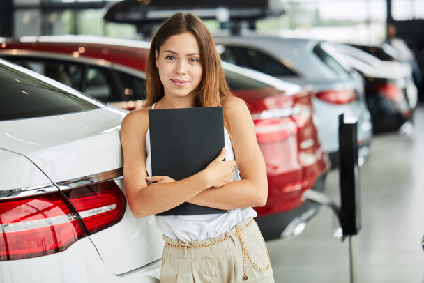 Female seller holding black folder looking at camera at car dealership. - Photo, image