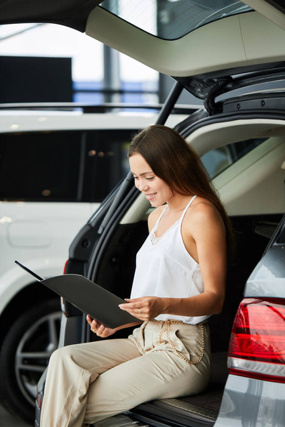 Smiling woman choosing new car at car showroom and reading properties - Photo, image