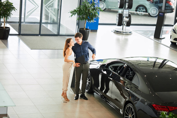 Young car salesman telling about features of car to female customer - Foto, Imagen