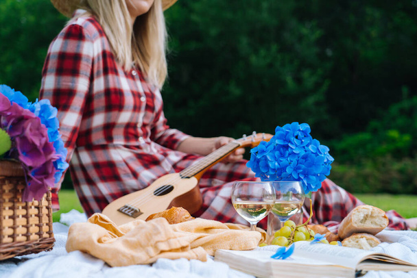 Chica en vestido rojo a cuadros y sombrero sentado en blanco punto manta de picnic juega ukelele y beber vino. Picnic de verano en el día soleado con pan, fruta, flores de hortensias ramo. Enfoque selectivo - Foto, Imagen