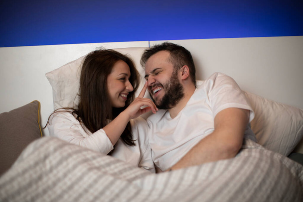Δύο εραστές στο κρεβάτι. Ευτυχισμένο κορίτσι Μπρουνέτ αγγίζοντας τη μύτη του γενειοφόρου φίλου - Φωτογραφία, εικόνα