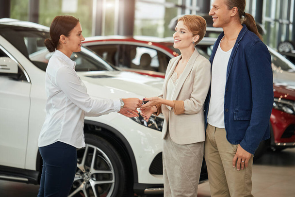 Happy young couple receiving car key from a salesman at the car dealership. - 写真・画像