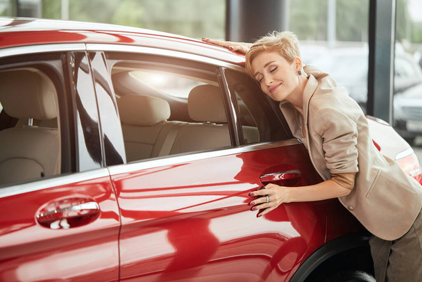 Happy caucasian woman embracing her new car at auto dealership - Foto, Imagen