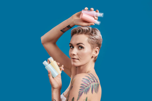 Portrait of beautiful tattooed woman with short hair looking at camera, holding plastic bottles with cosmetic, hair and skin care products isolated over blue background - Photo, image