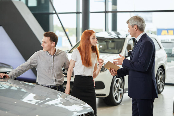 Car sales manager telling about the features of the car to the customers at the dealership - Фото, изображение