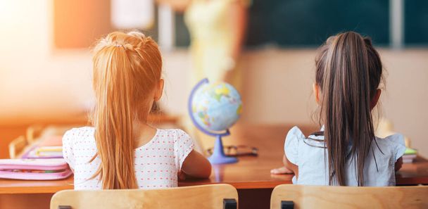 Dvě malé školačky sedí za stolem ve škole a pozorně poslouchají učitelku.. - Fotografie, Obrázek