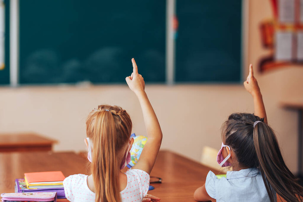 Zwei kleine Schulmädchen sitzen in einer Schulklasse am Schreibtisch und heben die Hände. - Foto, Bild