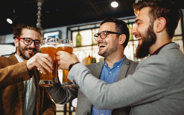 People, men, leisure, friendship and celebration concept. Happy business male friends drinking beer and clinking glasses at bar or pub - Fotó, kép