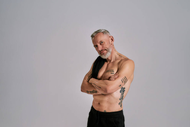 Middle aged muscular man took off black t shirt, showing his body, while posing in studio over grey background - Photo, image
