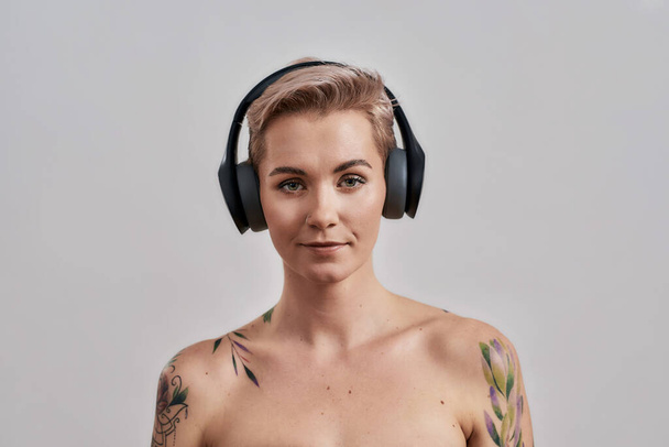 Attractive tattooed woman with pierced nose and short hair in headphones looking at camera while listening to music isolated over grey background - Fotó, kép