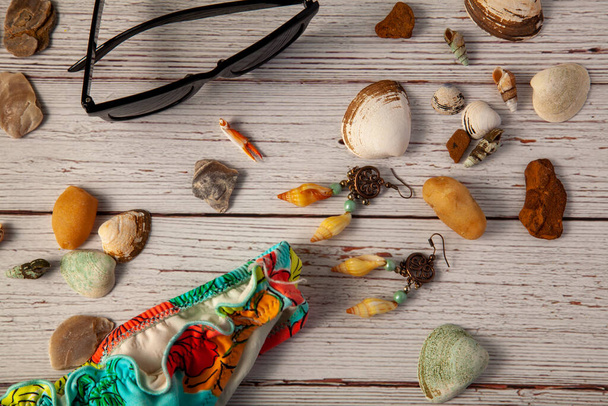 Een platte lay fotografie met een compositie van strand vakantie items, waaronder een zonnebril, een meisje zwembroek, schelpen, decoratieve stenen, oorbellen op houten achtergrond. - Foto, afbeelding