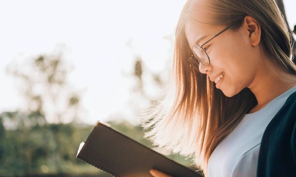 Donna sorridente in occhiali libro di lettura in parco all'aperto contro la luce del sole, concetto di stile di vita della città. - Foto, immagini