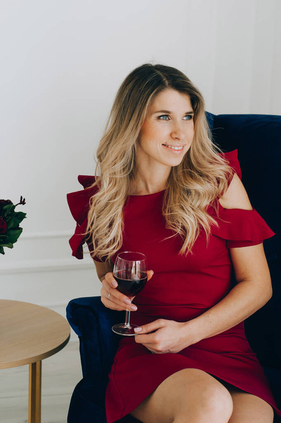 Portrait of a beautiful blonde girl in a red dress with a glass of red wine against the background of a stylish interior. - Фото, зображення