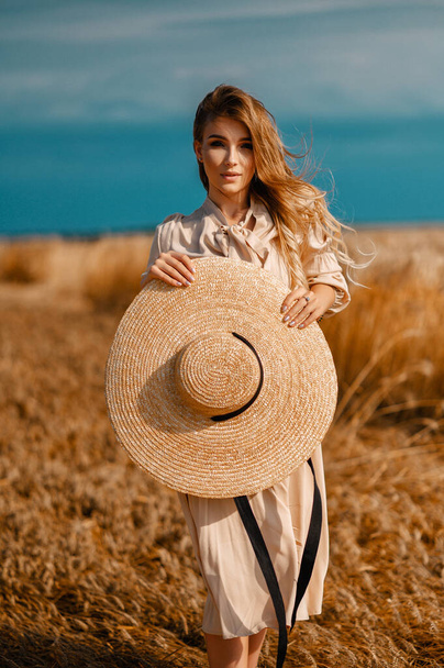 Attractive and beautiful girl in a beige midi dress with a big straw hat, in a wheat field - Fotó, kép