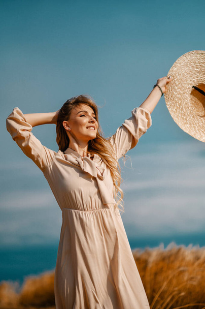 Attractive and beautiful girl in a beige midi dress with a big straw hat, in a wheat field - Zdjęcie, obraz