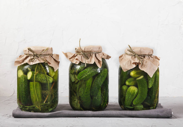 Three large glass jars with fermented cucumbers stand on a gray napkin against a concrete background. - Fotó, kép