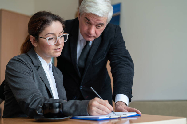 The male senior director watches as the female secretary signs the papers. Business woman fills out documents under the supervision of a boss in the office. - Photo, Image