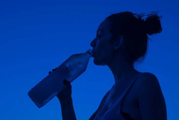 mladá dívka pití minerální vody z láhve s modrým pozadím - Fotografie, Obrázek
