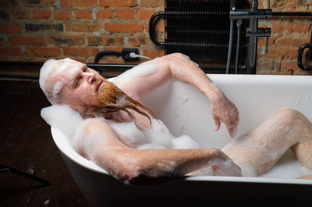 Close-up portrait of funny red-bearded bald man takes a bath with foam on the background of a brick wall. Cheerful naughty guy at spa treatments. A parody of glamorous girls. - Φωτογραφία, εικόνα