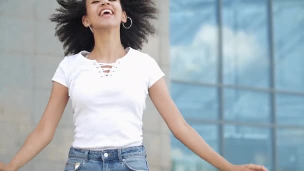 Cheerful excited afro millennial slender girl enjoys jumping on joyfully laughs smiling with wide open mouth waving hands turns keeping balance. Happy american young woman having fun enjoy movement. - Záběry, video