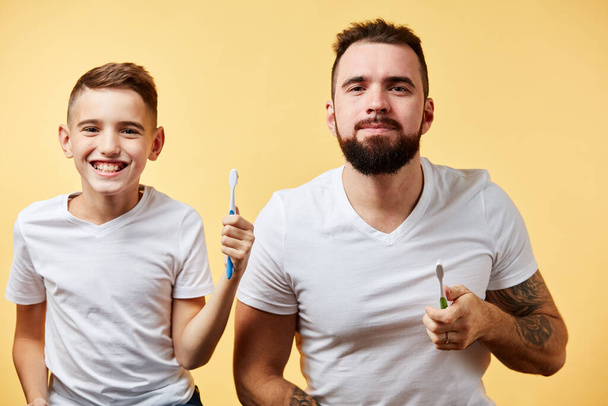 father and son brushing teeth and looking at camera together isolated on yellow - 写真・画像