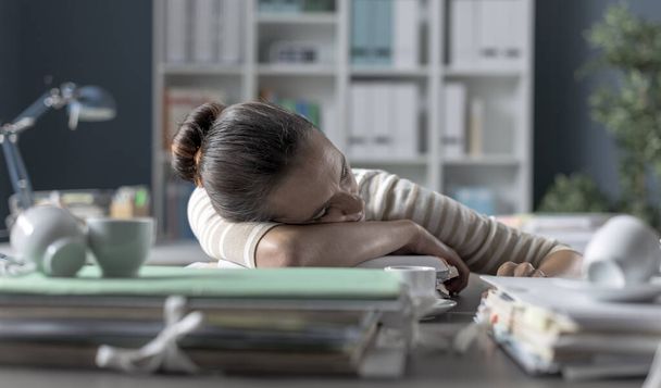Уставшая деловая женщина спит на столе, она устала и опирается на кучу бумажной работы - Фото, изображение